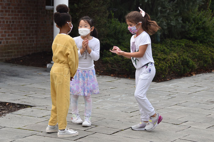 Three little girls playing in the schoolyard, from left a Black girl with a yellow clothes and her hair put up, a girl of Asian descent wearing a white top, pastel skirt, white tights and face mask, and a caucasian girl with a long brown onitail, white clothes and a patterned mask.