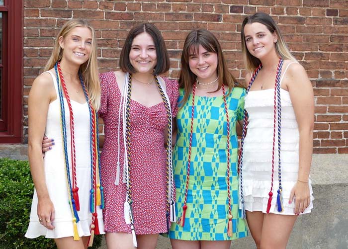 Four smiling female Elizabeth Morrow alumnae  wearing summer dresses and graduation tassels